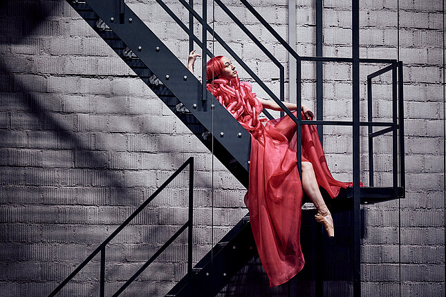 [Translate to Belgian French:] Une danseuse aux cheveux roux et en robe rouge pose sur un escalier en acier.