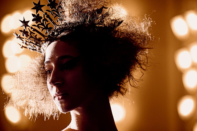[Translate to Luxembourgish:] Porträt einer Tänzerin mit kurzem, krausem Haar und einer Krone auf dem Kopf.