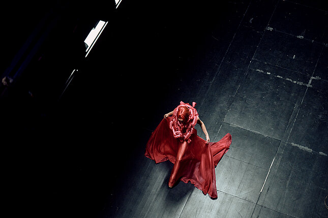 [Translate to Austrian:] Tänzerin mit rotem Haar und in rotem Kleid wandelt über die Bühne.