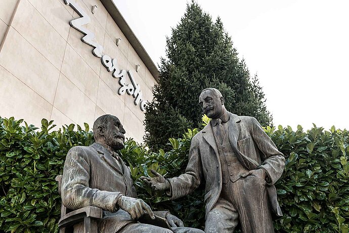 Deux hommes en costume, statues de bronze, devant un immeuble Marzotto.